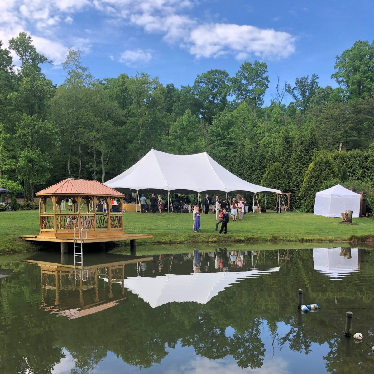 A white tent is set up for an event near a pond with a wooden gazebo on the grassy shore. Guests interact under clear skies with lush green trees in the background.