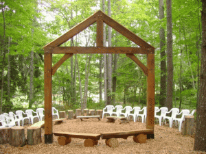 Outdoor ceremony setup with wooden arch, white chairs, and log seating arranged in a forest setting.