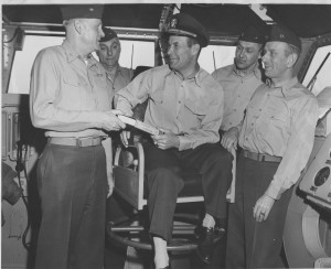 Five men in military uniforms are inside a control room, with one seated individual receiving a document from another standing man.