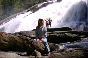 A person sits on a large rock in front of a waterfall, with two other people standing on rocks in the background.