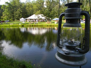 A black lantern hangs by a pond with a tent and people in the background surrounded by trees.