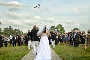 A wedding couple walks under a military arch of swords while guests look on. An aerial formation of planes flies overhead in the background.