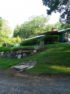 A green yard with a stone retaining wall leads up to a ranch-style house surrounded by trees and greenery. A small wooden bridge crosses a rocky feature in the foreground.