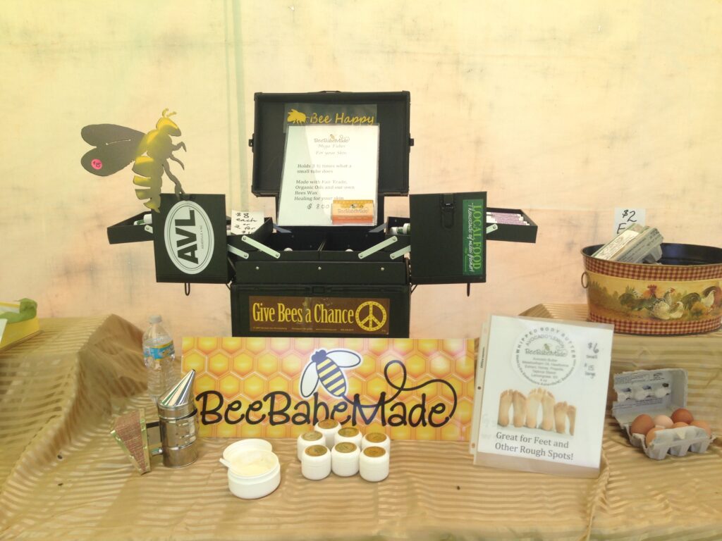 Display table featuring bee-themed skincare products, informational materials, and a donation box. Various items include creams, samples, and a promotional book titled "Great for Feet and Other Rough Spots.