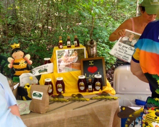 Outdoor market booth displaying honey products, including honey jars, honey sticks, and a honey bear plush. There are signs, a framed photo, wooden skewer props, and a person holding up a newspaper.