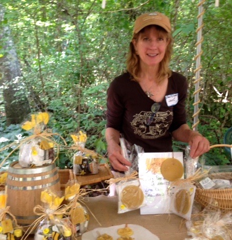 A woman stands behind a table with homemade goods, including jars and packaged items. She is smiling, wearing a brown shirt and a beige cap, and is in an outdoor, wooded setting.