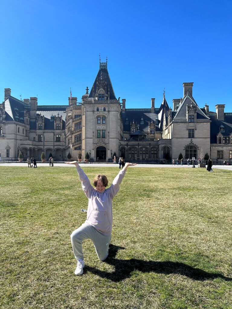 A person in casual clothes poses with raised arms in front of a large, historic mansion on a sunny day.
