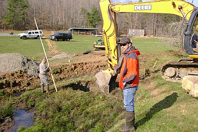 Two workers in a field stand near a trench that is being excavated by a yellow Deere excavator. One worker measures the trench depth with a leveling rod. Vehicles and trees are in the background.