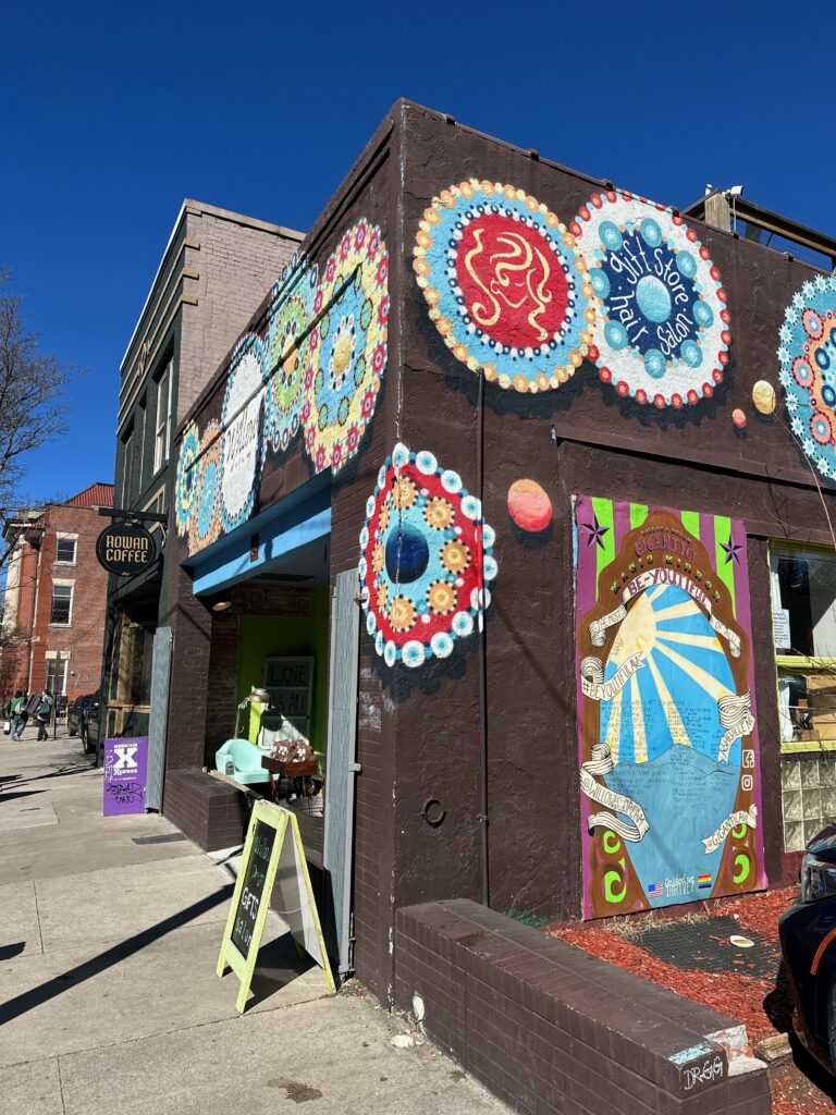 Colorful mural covers the exterior of a building housing "Round About Coffee." The scene includes vibrant designs and a sign outside advertising iced coffee. A sunny day with clear blue skies.