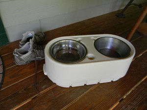 A pair of hiking boots is placed next to an elevated pet feeding station with two stainless steel bowls on a wooden floor.