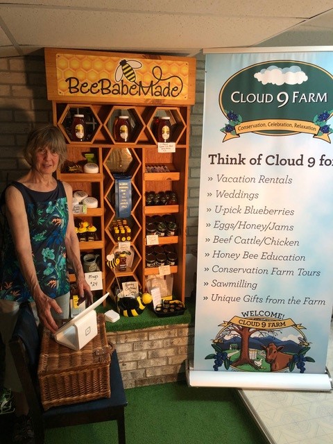 A woman stands beside a display of honey products labeled "Bee Babe Made" and a Cloud 9 Farm banner listing various farm activities and products.